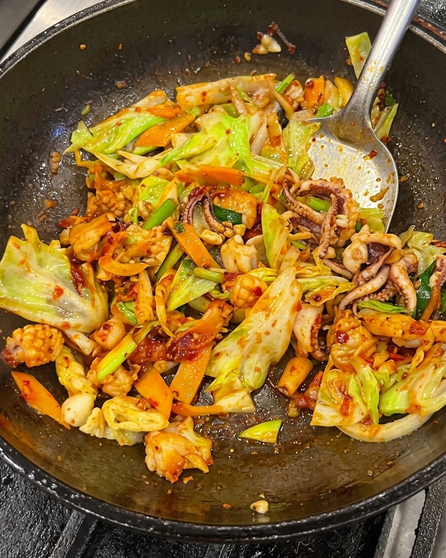 Primer plano de calamar, panceta, repollo y zanahorias siendo salteados con una espátula de metal dentro de un wok, completamente cubiertos de una salsa picante roja y brillante.