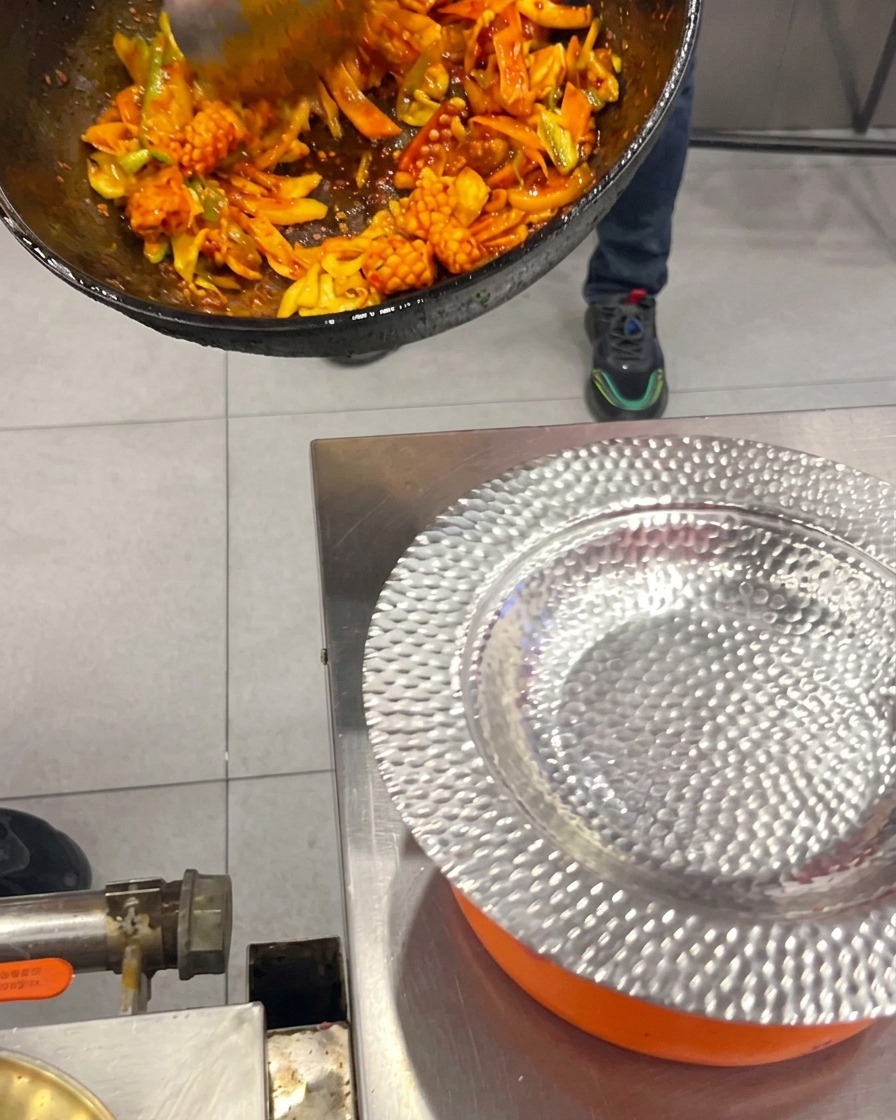 Salteado picante coreano de calamar y panceta con verduras siendo servido desde un wok hacia un plato de plata labrada.
