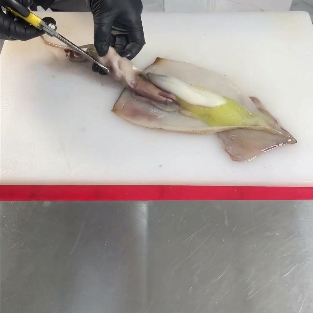 Manos con guantes usando tijeras para abrir y limpiar un calamar crudo sobre una tabla de cortar blanca.