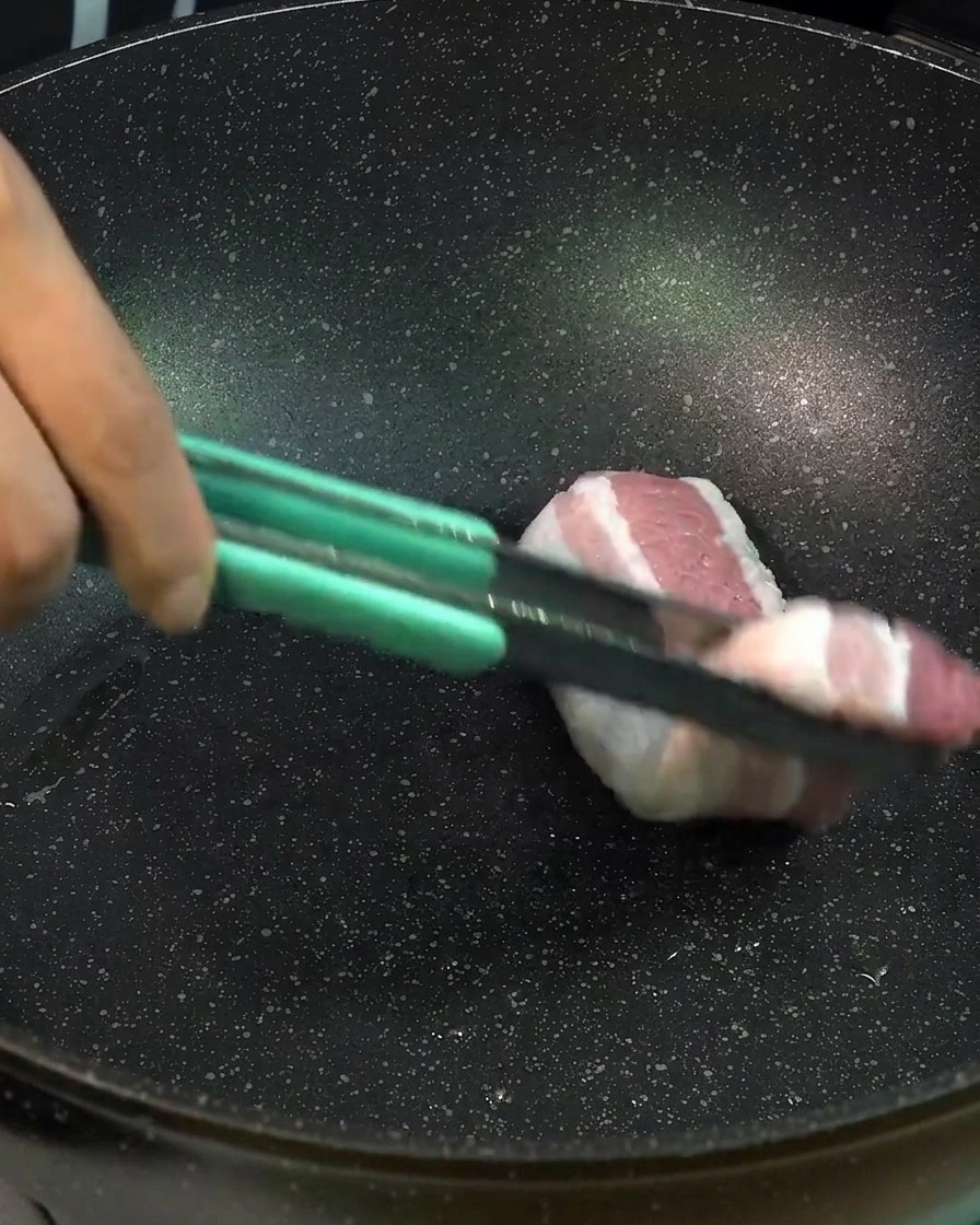 Rodajas gruesas de panceta de cerdo cruda colocándose en una sartén negra con unas pinzas de color verde claro para dorar la carne.