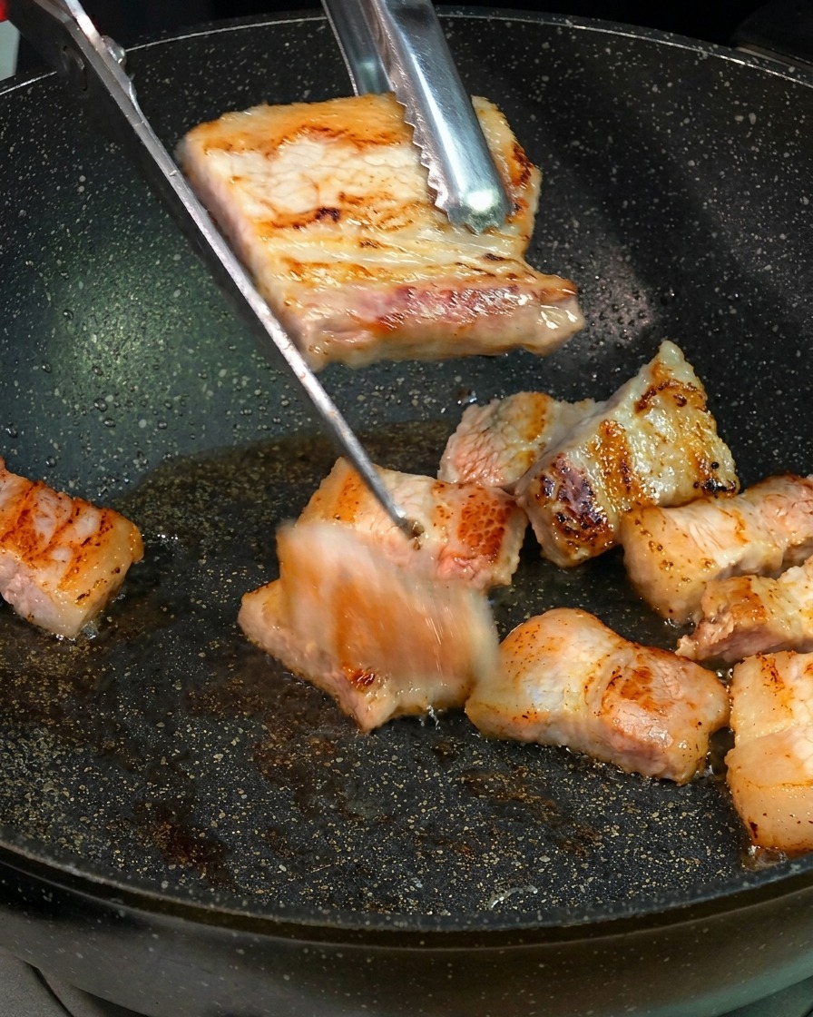 Rodajas de panceta de cerdo doradas cortándose en trozos más pequeños con tijeras de cocina sobre una sartén.
