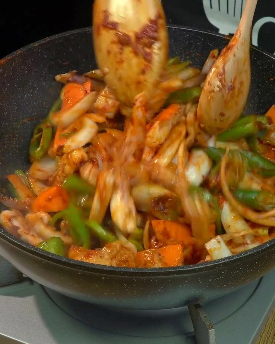 Efecto de movimiento de panceta, calamar y verduras mezcladas siendo salteados vigorosamente con dos cucharas de madera en un wok, completamente cubiertos de salsa picante roja.