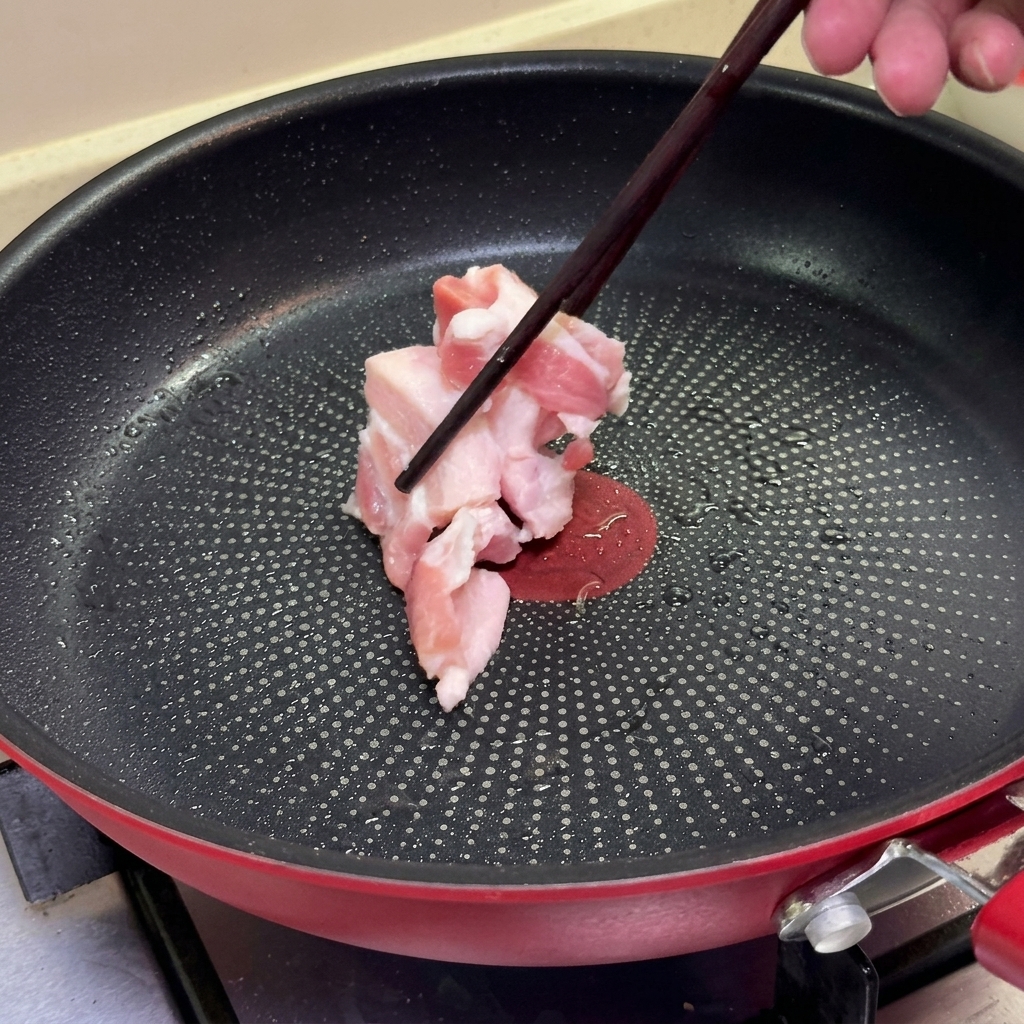 Unos palillos colocando rodajas de panceta de cerdo cruda en una sartén antiadherente caliente.