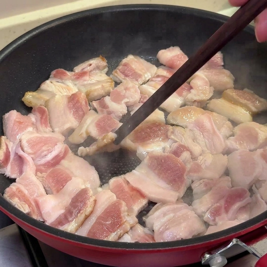 Tiras de panceta de cerdo friéndose en una sartén antiadherente, dorándose y caramelizándose ligeramente mientras se remueven con palillos.