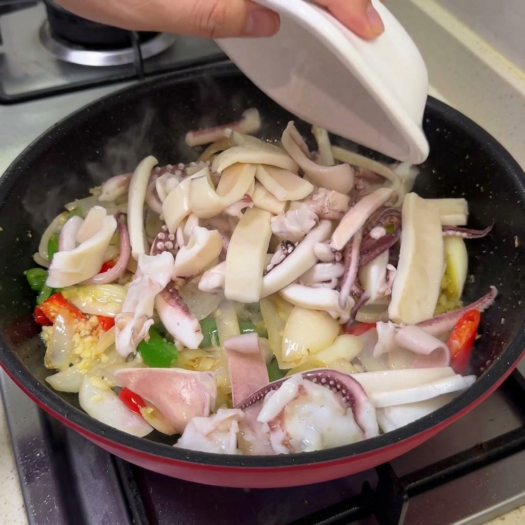 Trozos de calamar escaldado y panceta de cerdo dorada vertiéndose desde un tazón a una sartén con verduras salteadas.