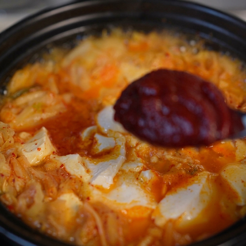 Una cuchara sosteniendo una porción de pasta de chile coreana, espesa y roja, sobre la sopa de kimchi y tofu hirviendo.