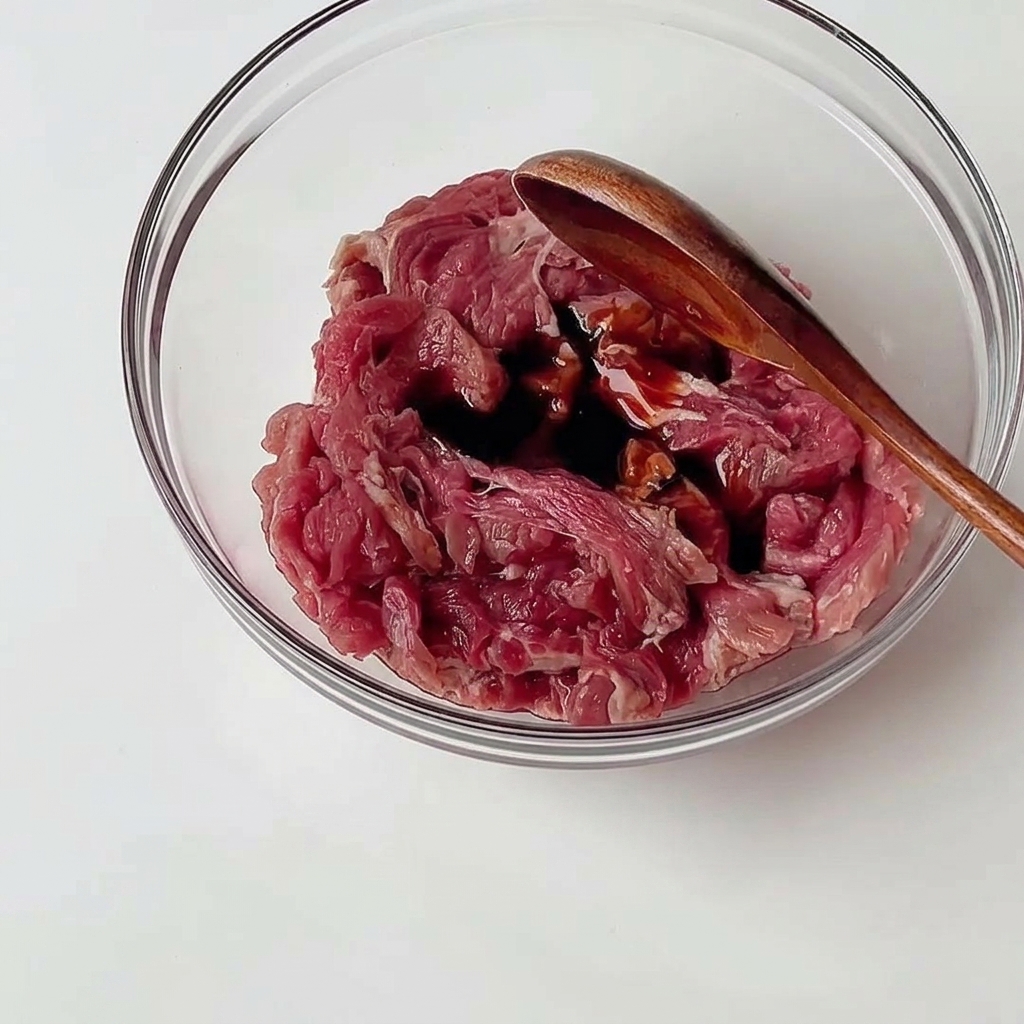 Tiras finas de ternera cruda en un bol de vidrio mezclándose con salsas oscuras usando una cuchara de madera.