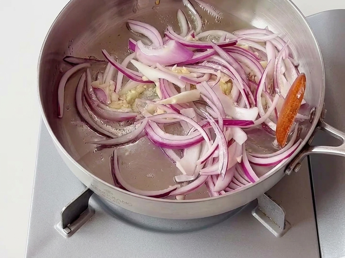 Rodajas de cebolla morada y trozos de ajo picado sofriéndose en una sartén de acero inoxidable sobre una estufa portátil.