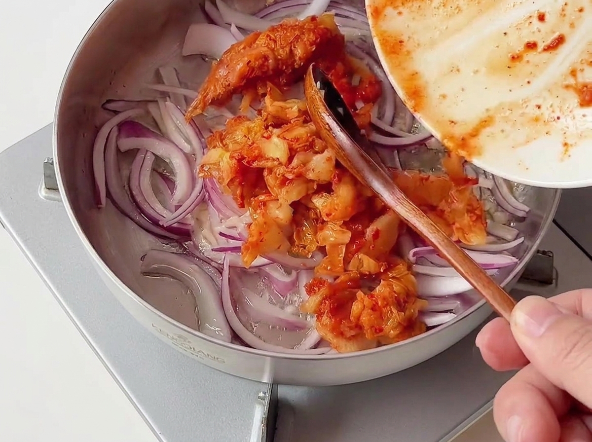 Una porción de kimchi rojo picado añadiéndose desde un plato a una sartén de acero inoxidable con cebolla morada.