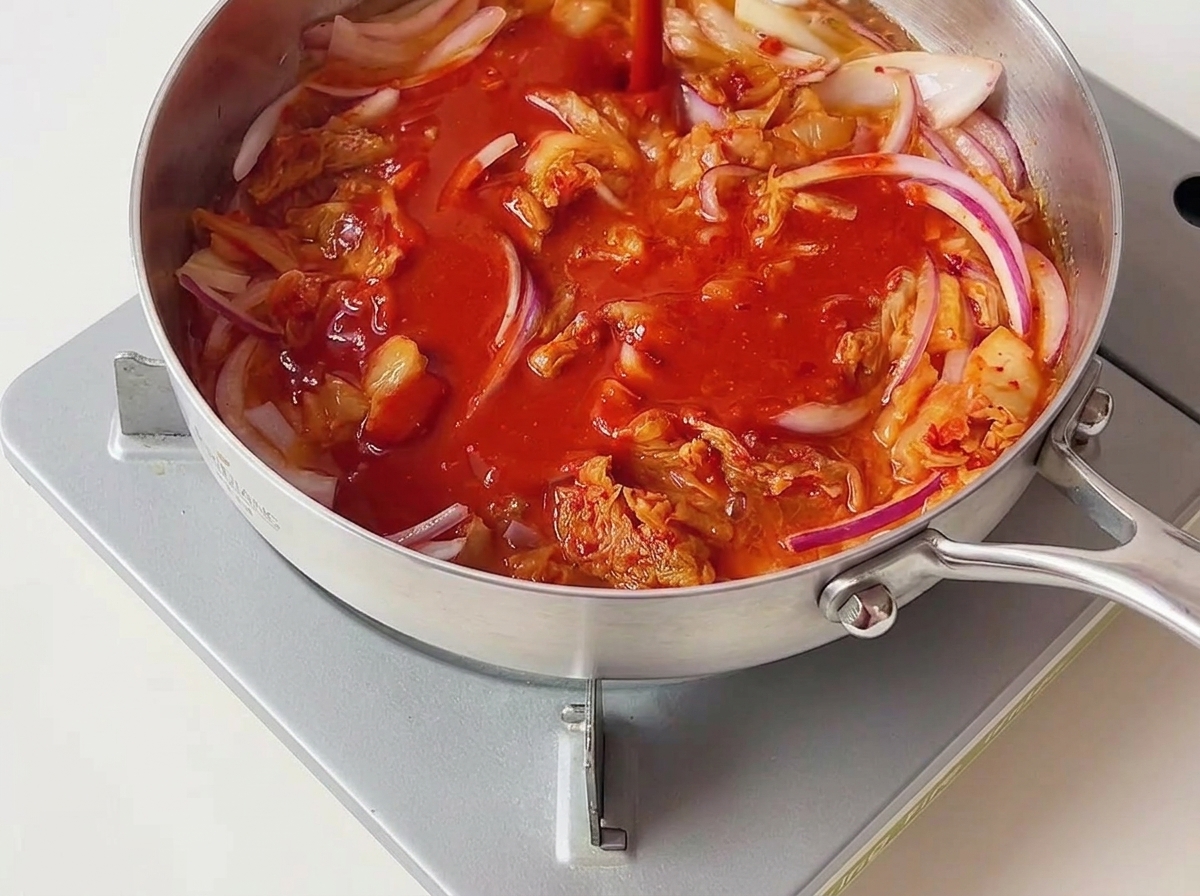 Salsa de chile de color rojo brillante vertiéndose sobre el kimchi y la cebolla salteados en una sartén plateada.
