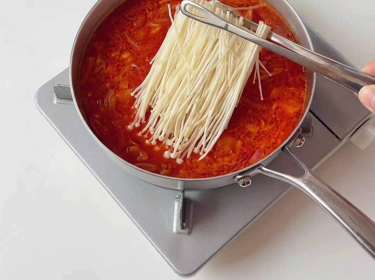 Unas pinzas metálicas colocando un manojo de setas enoki blancas en un estofado de kimchi rojo que burbujea.