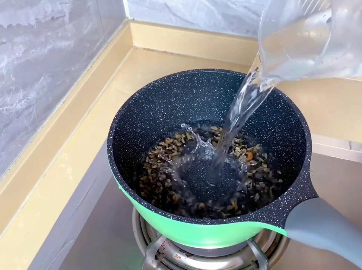Agua limpia vertiéndose desde una jarra medidora de plástico en una olla verde sobre una estufa de gas.