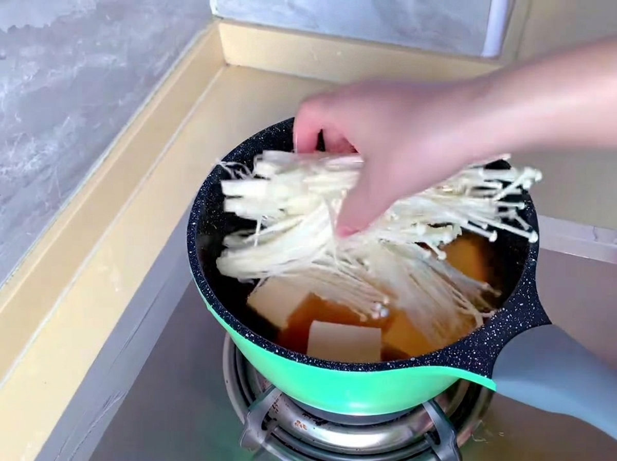 Una mano transfiere un manojo de setas enoki blancas y alargadas a una olla que ya contiene caldo y tofu.