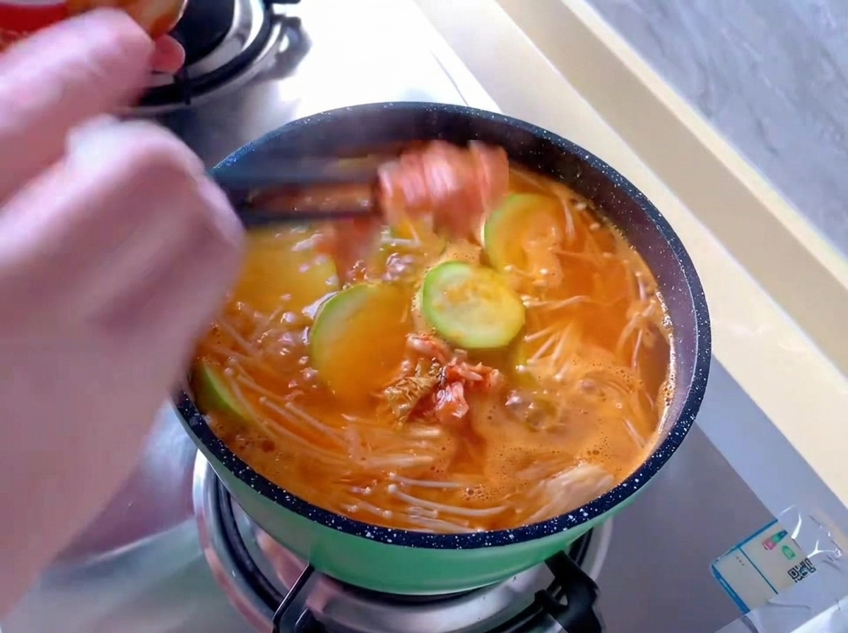 Añadiendo trozos de col kimchi roja con palillos en una pequeña olla verde de sopa naranja hirviendo con setas enoki y rodajas de calabacín.