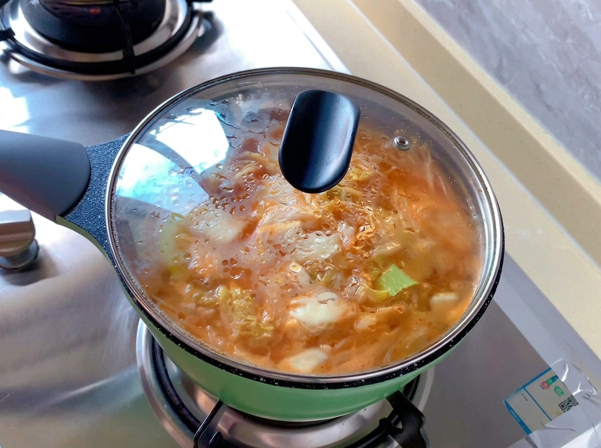 Una pequeña olla verde llena de sopa de kimchi naranja tapada con una tapa de vidrio transparente con condensación de vapor en la superficie.
