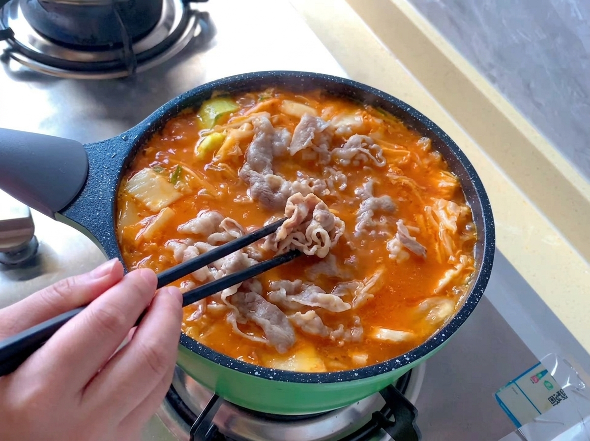 Rollos de ternera blanqueados distribuyéndose con palillos negros dentro de una olla hirviendo de sopa de kimchi de color rojo anaranjado brillante.