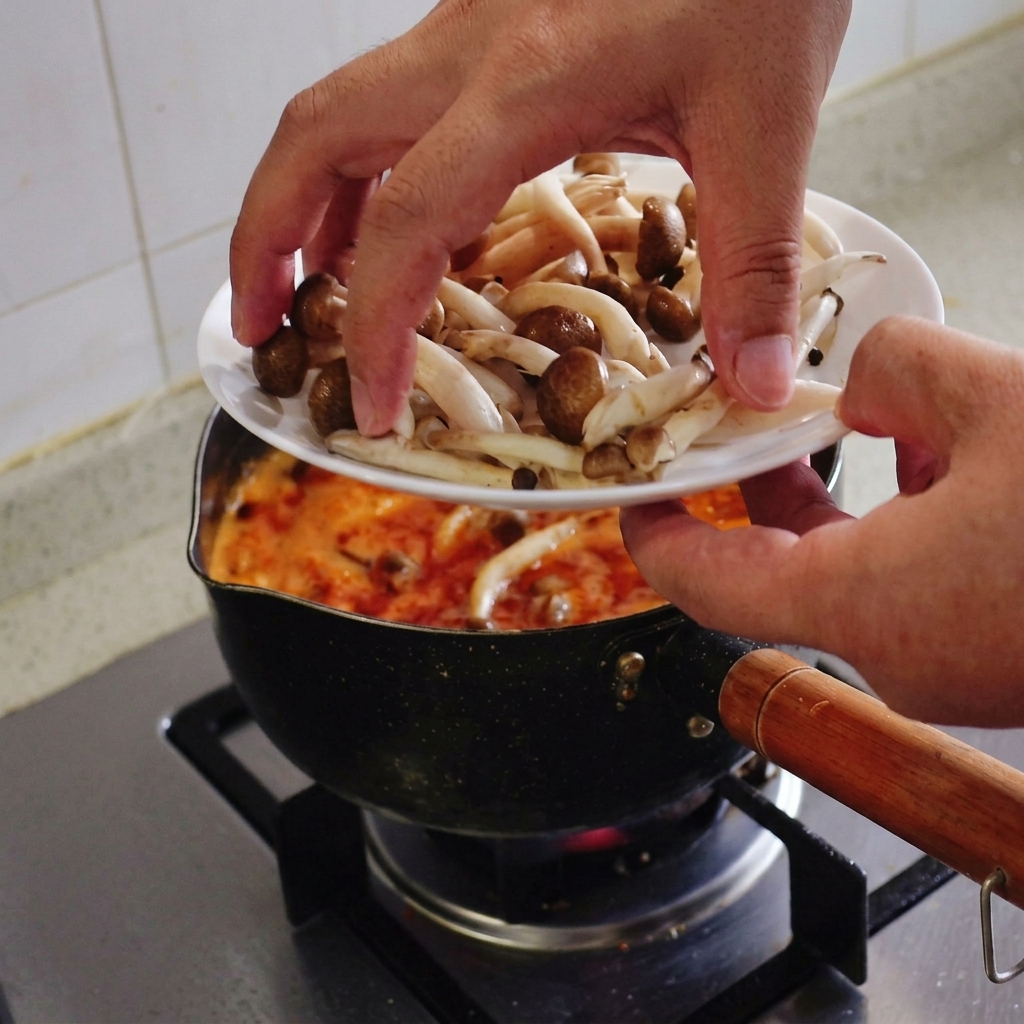 Unas manos pasando un puñado de setas shimeji frescas con sombrerito marrón desde un plato hacia la sopa hirviendo.