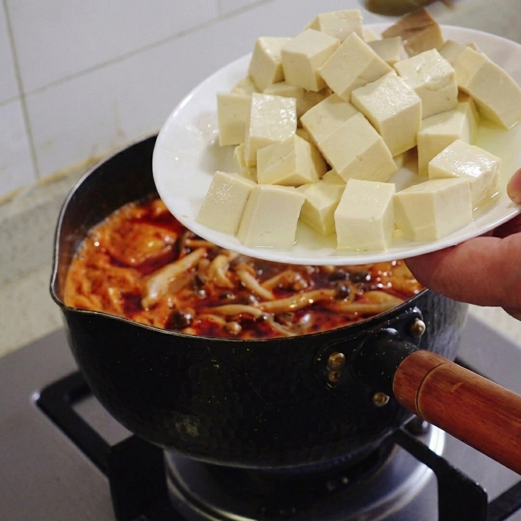 Una mano sosteniendo un plato blanco y deslizando cubos perfectos de tofu suave en el estofado de kimchi.