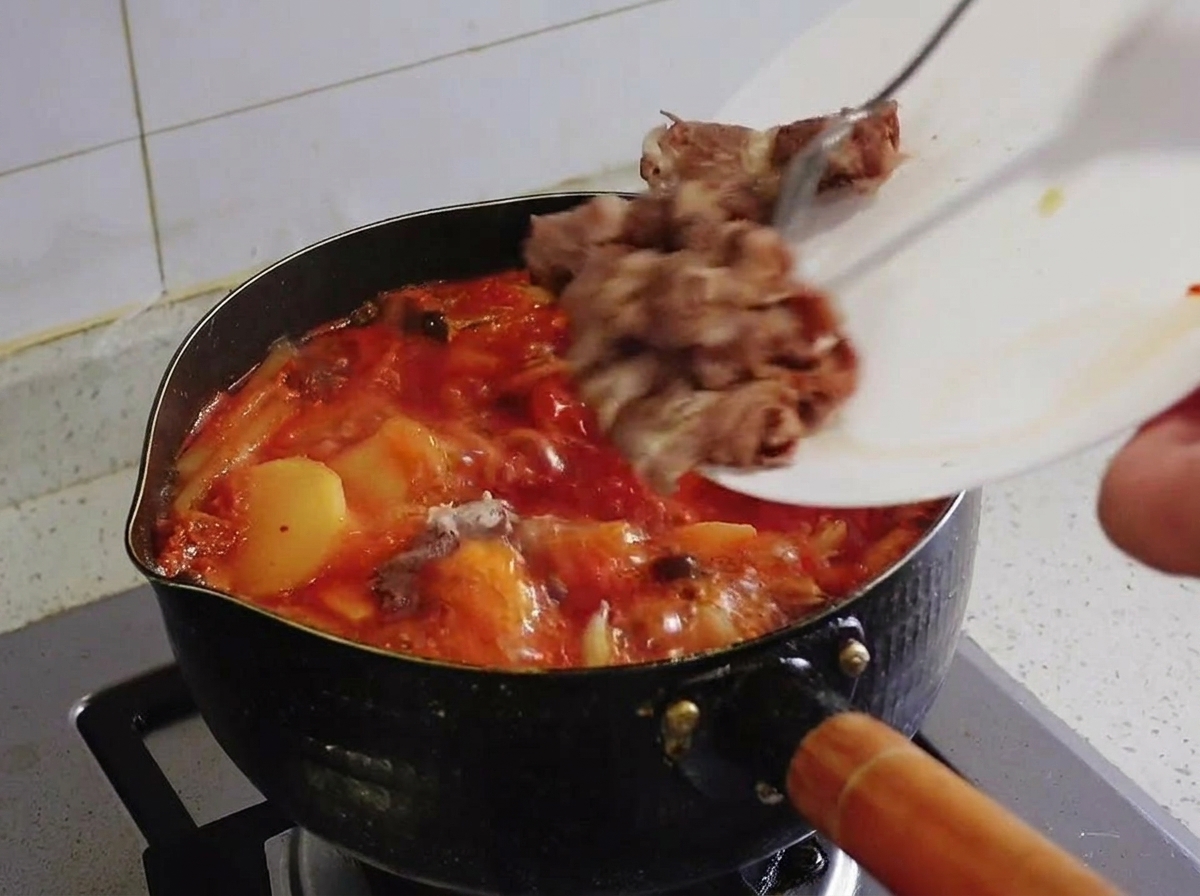 Trozos de ternera veteada blanqueada deslizándose desde un plato blanco hacia el estofado de kimchi hirviendo.