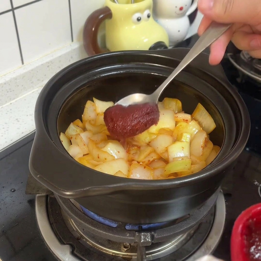 Una cuchara de metal mezclando pasta de chile coreana roja y espesa con el kimchi y las cebollas sofritas dentro de la olla de barro.