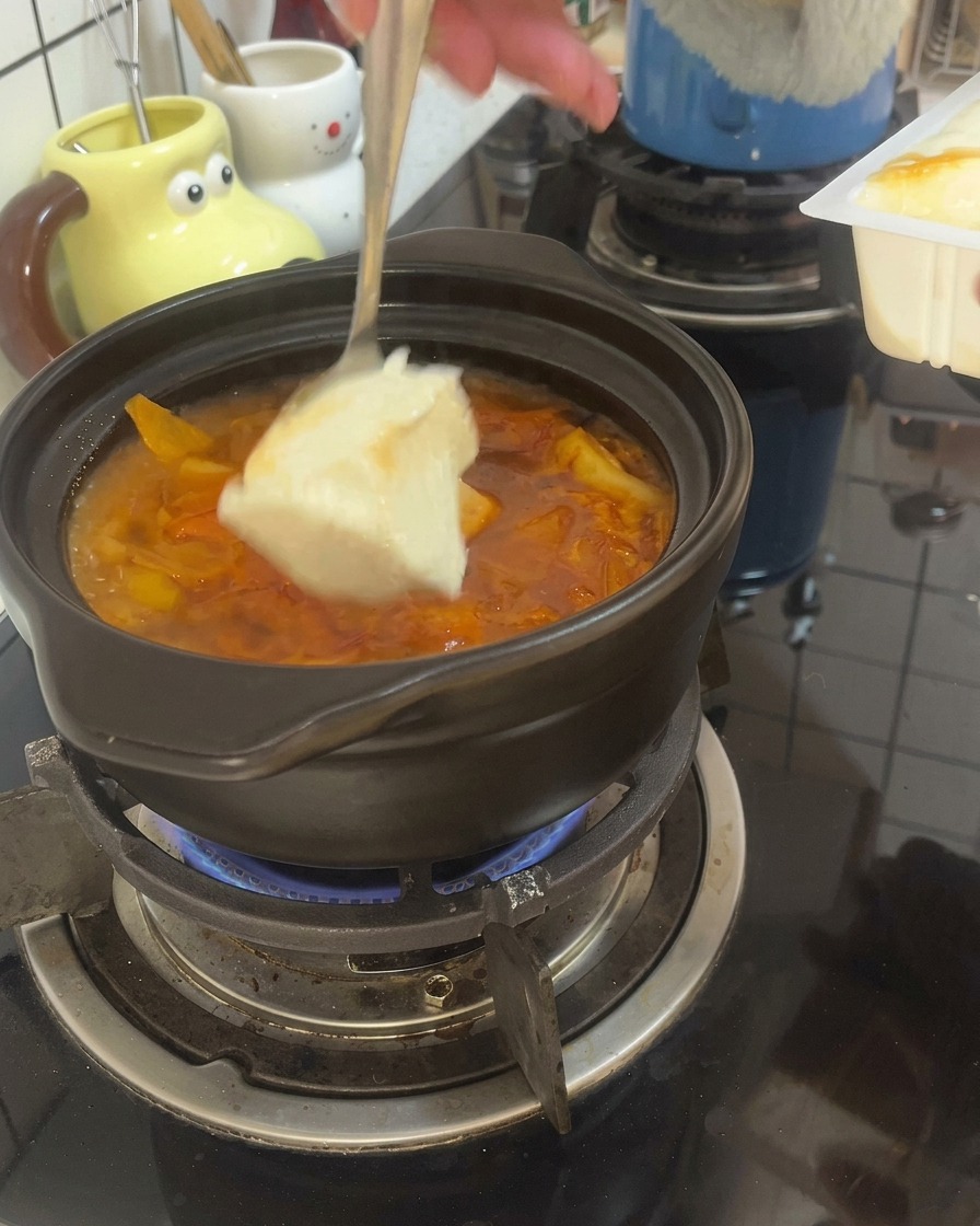 Una cuchara sirviendo trozos grandes de tofu suave en un caldo rojo burbujeante dentro de una olla de barro.