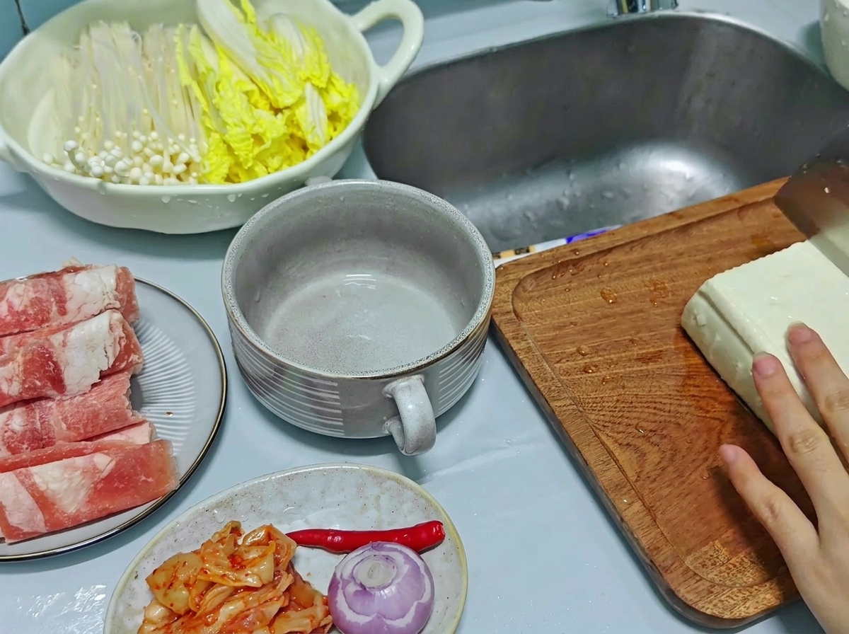 Un primer plano de una mano usando un cuchillo de cocina para cortar un bloque grande de tofu blanco en rodajas rectangulares gruesas sobre una tabla de madera.
