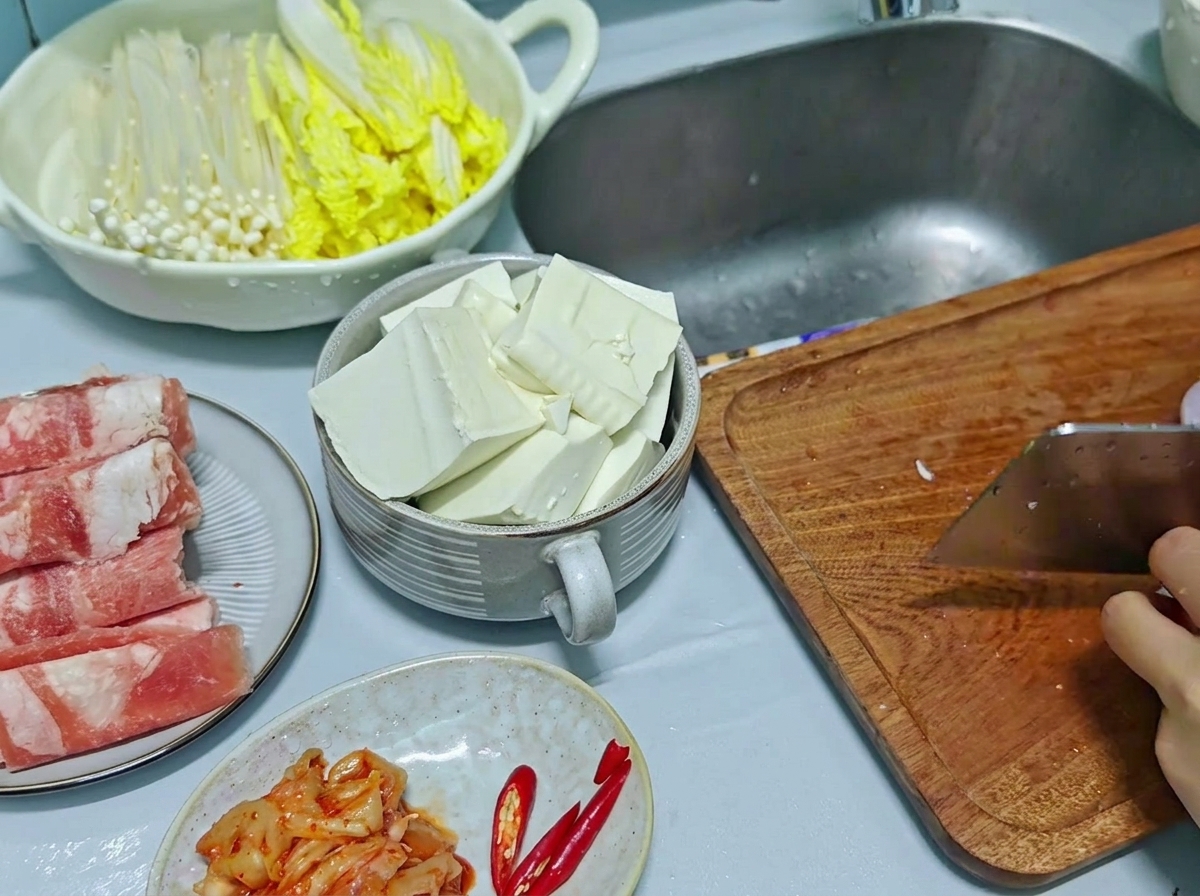 Un cuchillo de cocina picando cebolla roja y chiles rojos en una tabla de cortar de madera, con tofu cortado visible en un bol a un lado.
