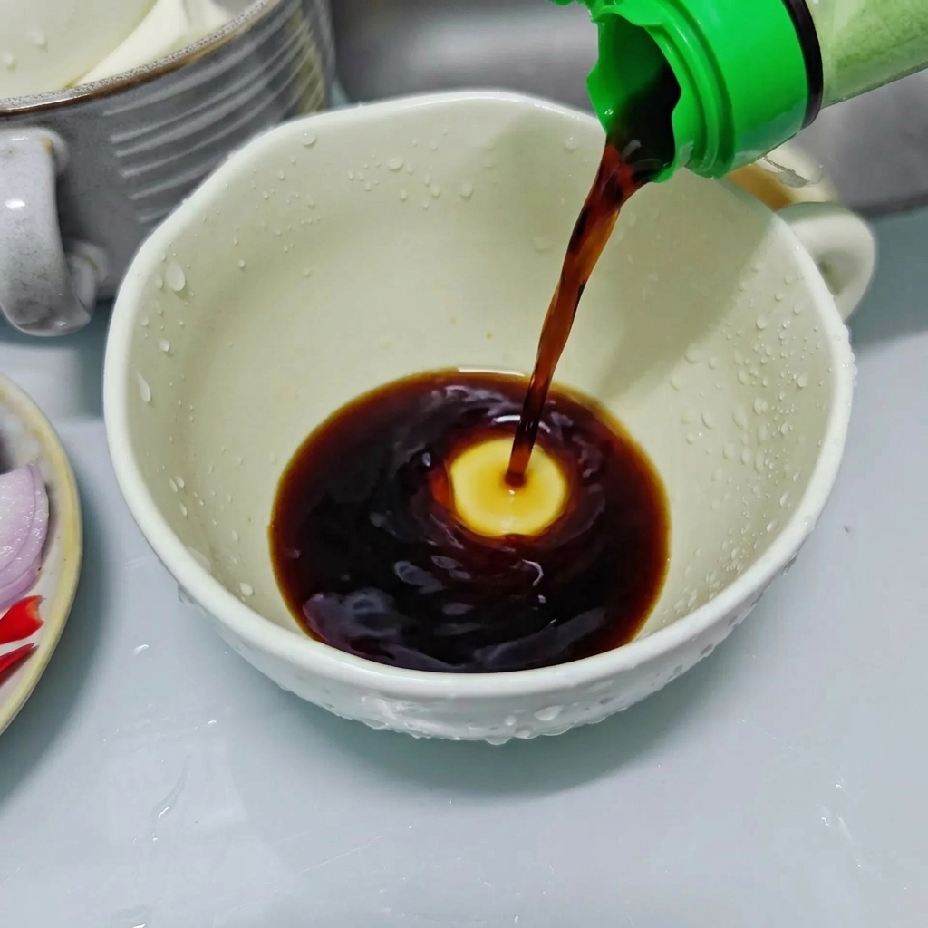 Salsa de soja oscura vertiéndose desde una botella de tapa verde en una taza de cerámica blanca para preparar el condimento.