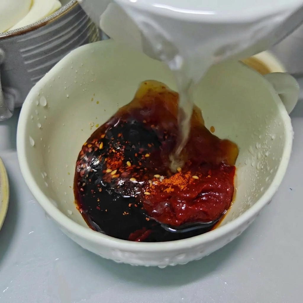 Agua clara vertiéndose en un tazón blanco lleno de salsa de soja oscura, pasta de chile rojo y hojuelas de chile.