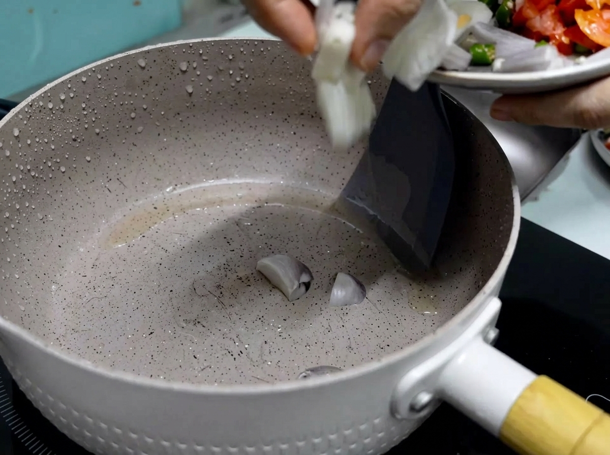Una mano deslizando cebollas rojas recién cortadas desde un plato pequeño a una sartén antiadherente caliente con aceite.