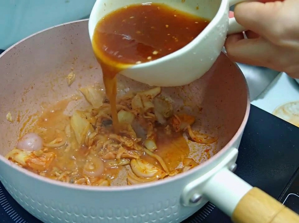 Una persona vertiendo una salsa rojiza mezclada desde una taza blanca a una sartén rosa que contiene kimchi y cebolla sofreída.