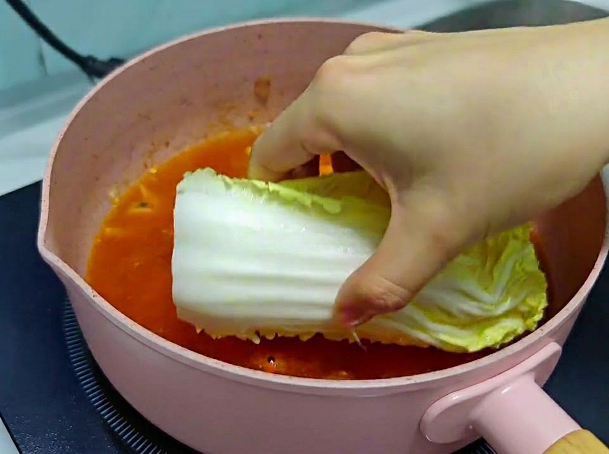 Una mano colocando cuidadosamente un trozo grande de col china fresca en una sartén rosa llena de caldo burbujeante.