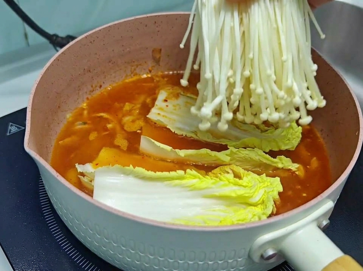 Un primer plano de una mano bajando un manojo fresco y limpio de setas enoki blancas en una olla de cocina rosa que contiene caldo de kimchi y col china.