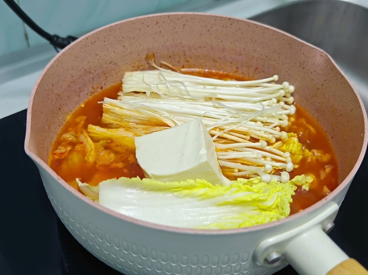 Un bloque grande de tofu blanco fresco descansando sobre setas enoki y repollo dentro de una olla de sopa de kimchi naranja.