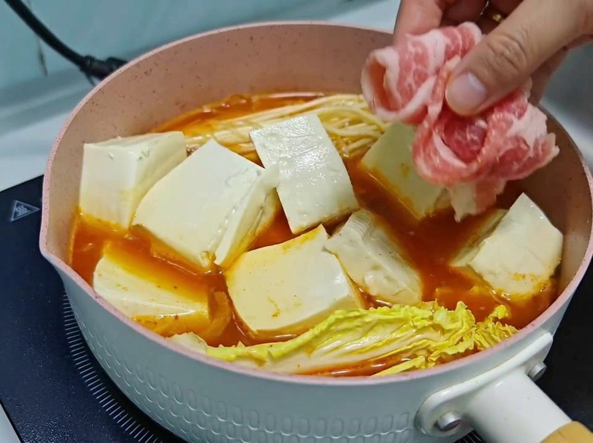 Una mano acomodando con cuidado finos rollitos de carne cruda sobre cubos de tofu y verduras en una olla de sopa de kimchi a fuego lento.