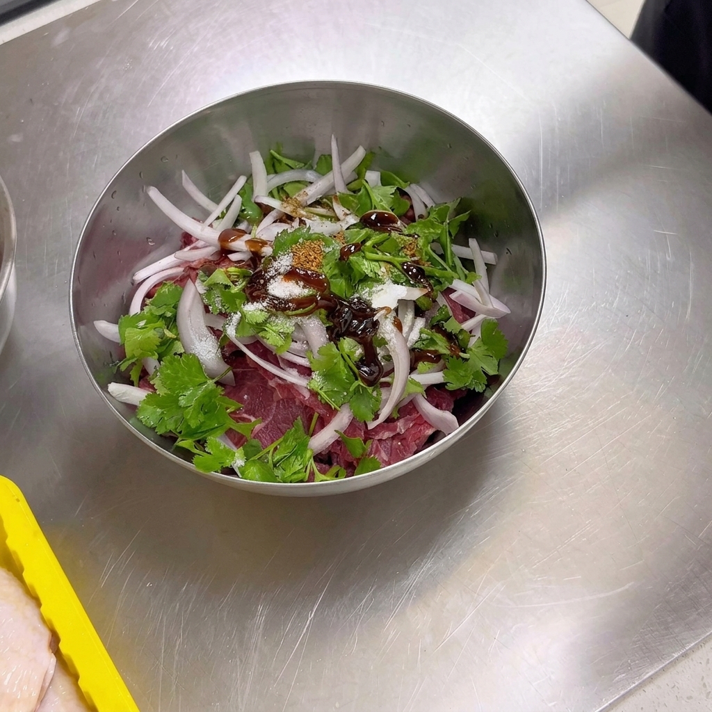 Un bol de metal que contiene ternera cruda laminada, cebolla y cilantro, cubierto con especias secas que incluyen comino en polvo, semillas de comino, sal y azúcar.