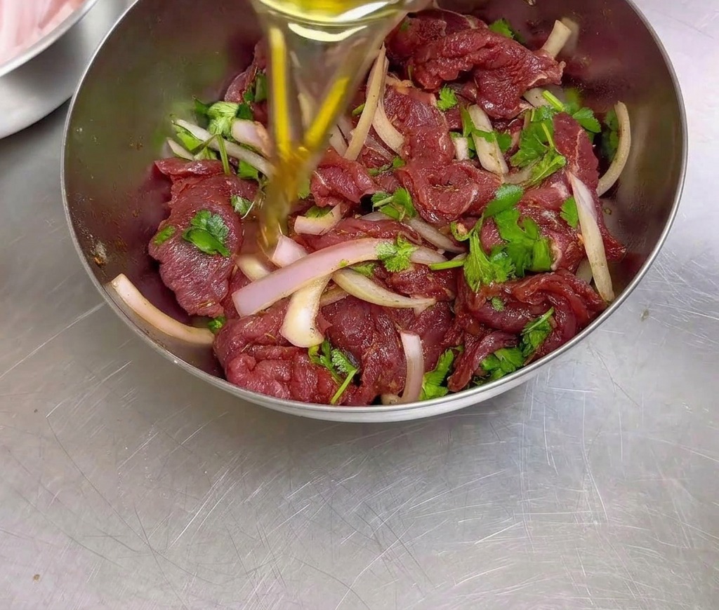 Una persona vierte aceite de cocina en un bol de acero inoxidable lleno de ternera sazonada, cebolla laminada y cilantro.