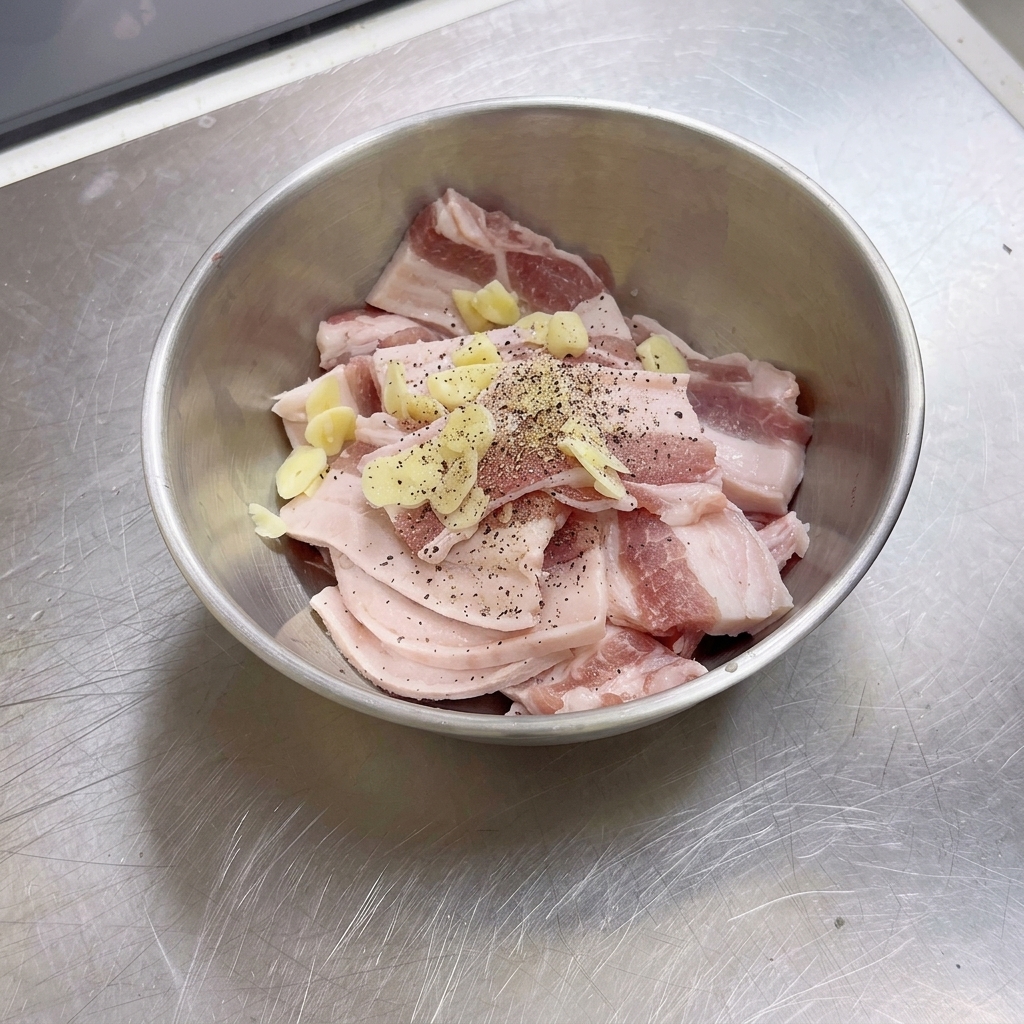 Un bol de acero inoxidable lleno de láminas de panceta cruda, trozos de ajo y pimienta negra recién molida.