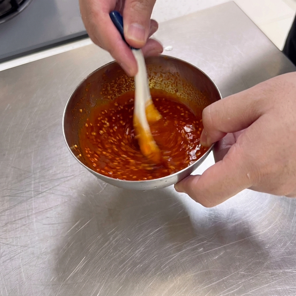 Una salsa espesa de color rojo oscuro para barbacoa con semillas de sésamo se remueve con una cuchara en un pequeño bol de acero inoxidable.