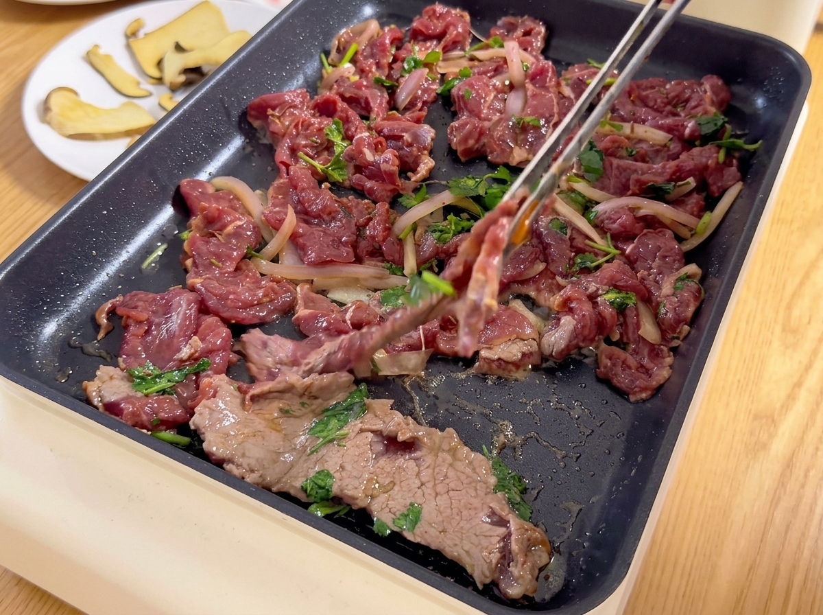 Ternera marinada cortada fina y mezclada con cebolla y cilantro se coloca sobre una parrilla eléctrica caliente con unas pinzas.