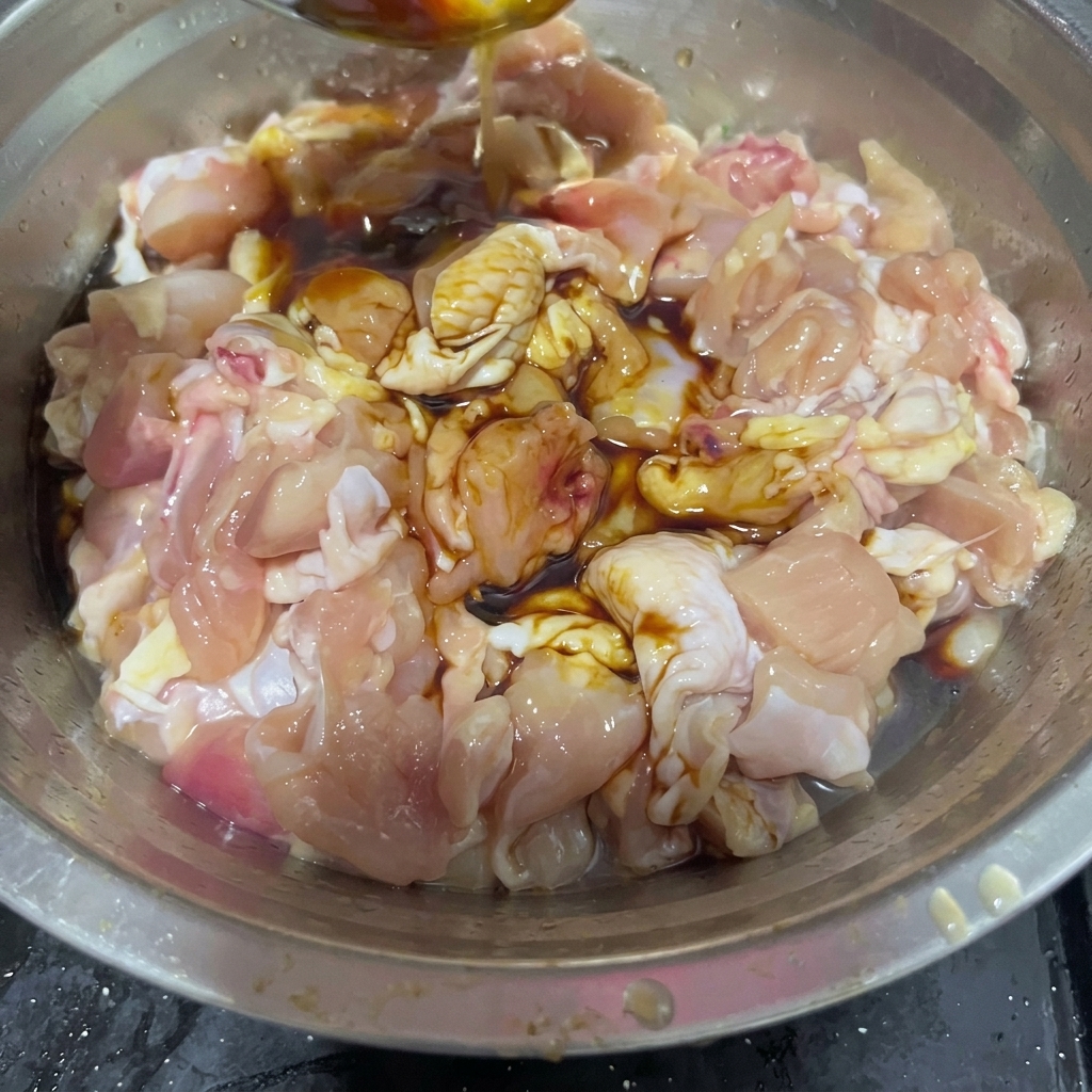 Vino de cocina vertiéndose desde una cuchara de acero inoxidable en un tazón que contiene trozos de pollo crudo y salsas oscuras.