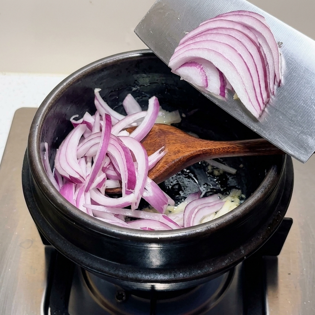 Un montón de cebolla morada rebanada pasándose desde la hoja de un cuchillo picador directamente a una olla negra con ingredientes aromáticos.