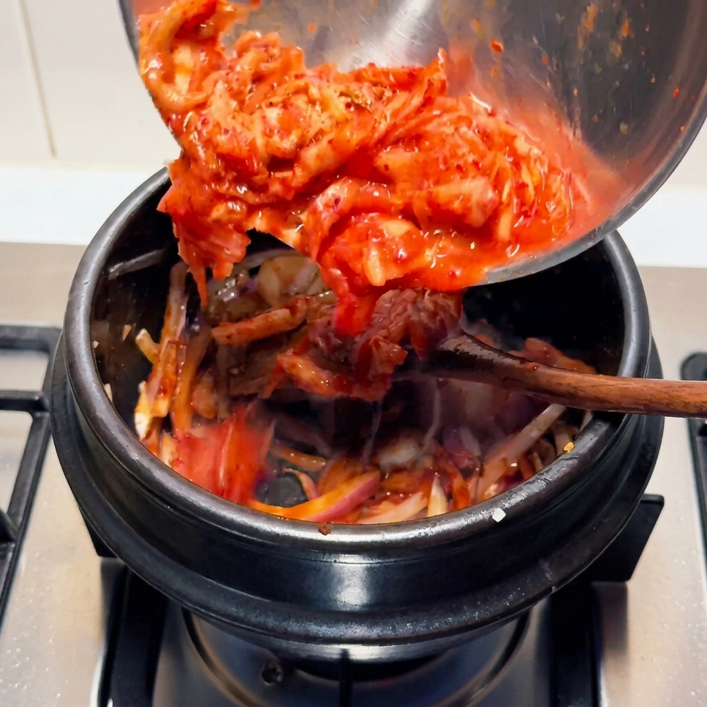 Kimchi agrio picado de color rojo brillante vertiéndose desde un tazón de acero inoxidable a una olla de piedra negra en la estufa.