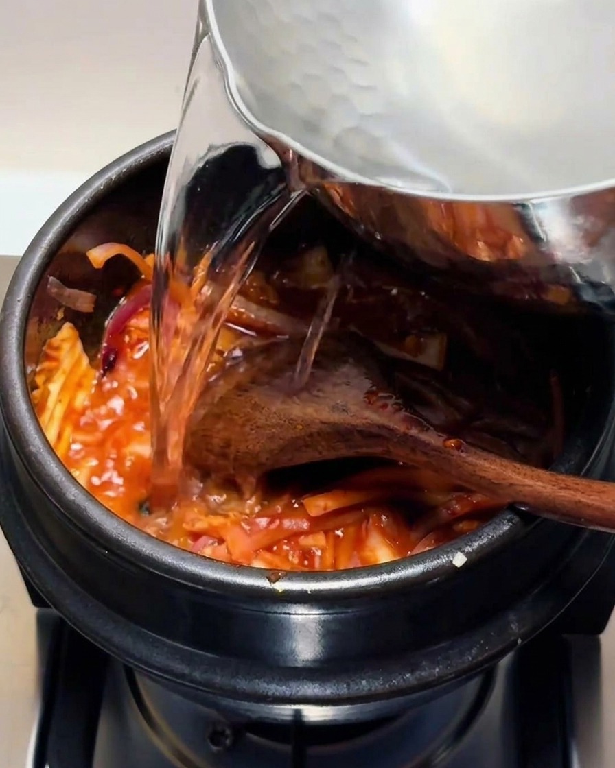 Agua caliente y transparente vertiéndose desde una olla de metal a una olla de piedra negra que contiene la mezcla de kimchi rojo.
