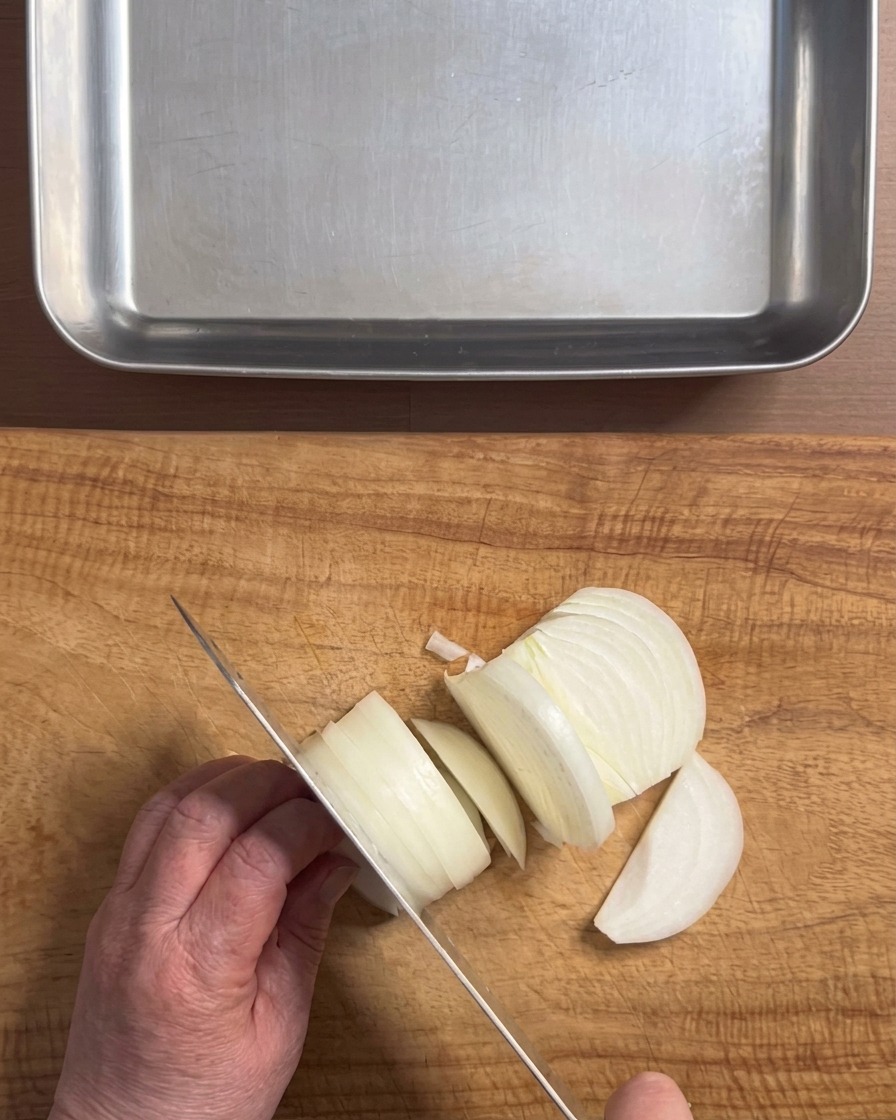 Manos usando un cuchillo para cortar una cebolla blanca en tiras gruesas sobre una tabla de cortar de madera.