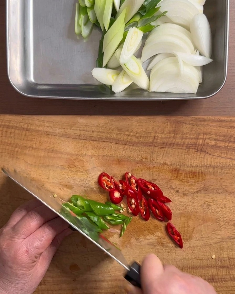 Manos usando un cuchillo para picar chiles verdes y rojos en rodajas sobre una tabla de cortar de madera.