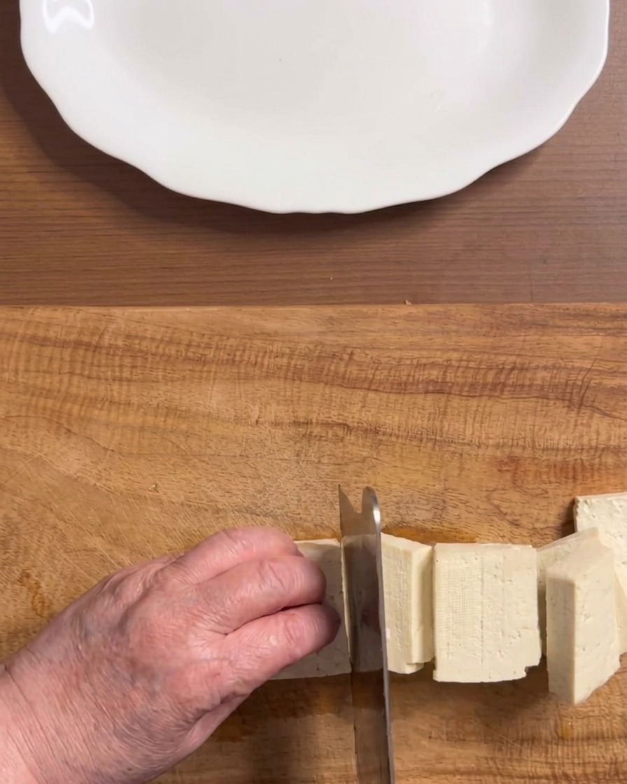 Manos cortando un bloque de tofu blanco firme en trozos rectangulares uniformes sobre una tabla de madera.