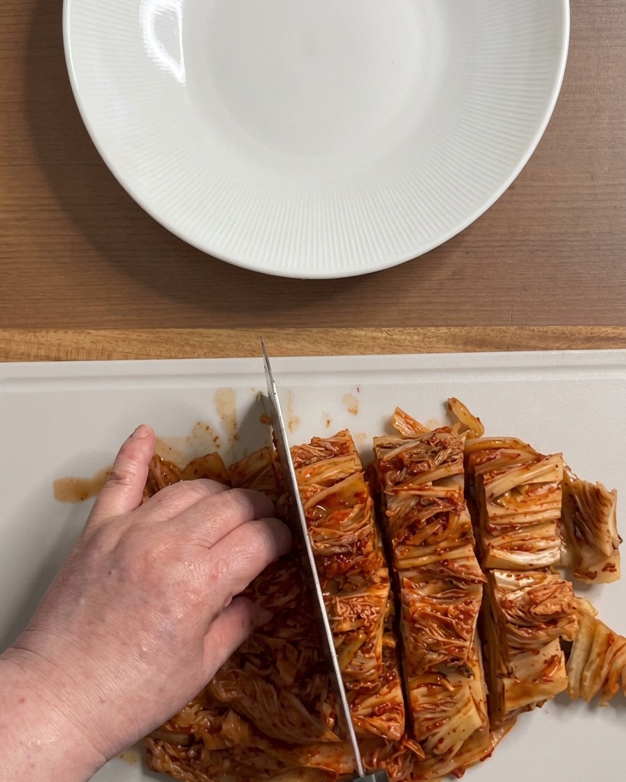 Manos cortando un bloque grande de kimchi rojo vibrante en trozos medianos sobre una tabla de cortar blanca.