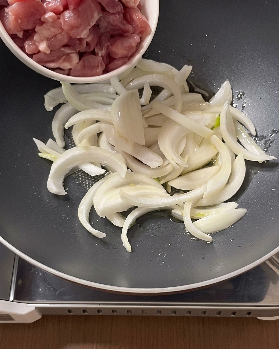 Trozos de carne de cerdo cruda siendo añadidos desde un cuenco pequeño a una olla con cebolla laminada y aceite.