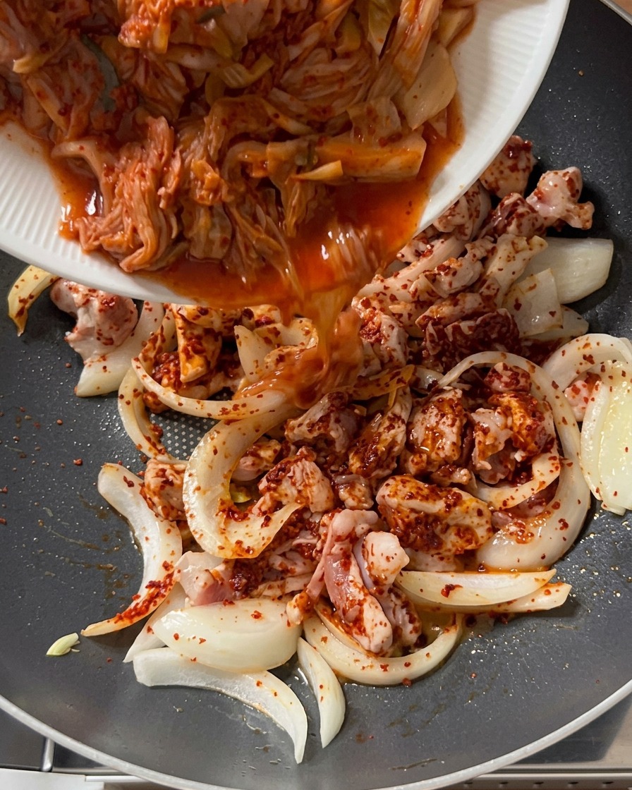 Kimchi troceado siendo vertido desde un plato blanco a una olla que contiene cerdo sazonado y cebollas.